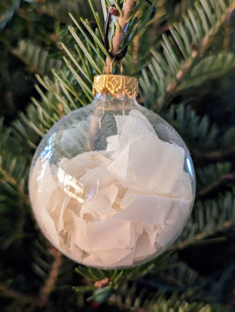 A clear glass Christmas ornament filled with ivory organza fabric from a mother's 1945 wedding dress hanging on a Christmas tree, a beautiful symbol of healing grief after losing your mother at the holidays