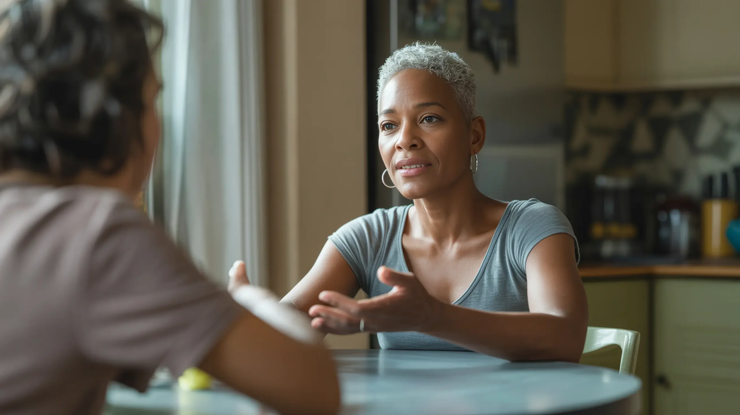 A Black woman in her 50s with natural gray hair sits at a kitchen table in open, relaxed conversation — an image of the ease and honesty that comes with saying "I don't know" as a sign of strength.