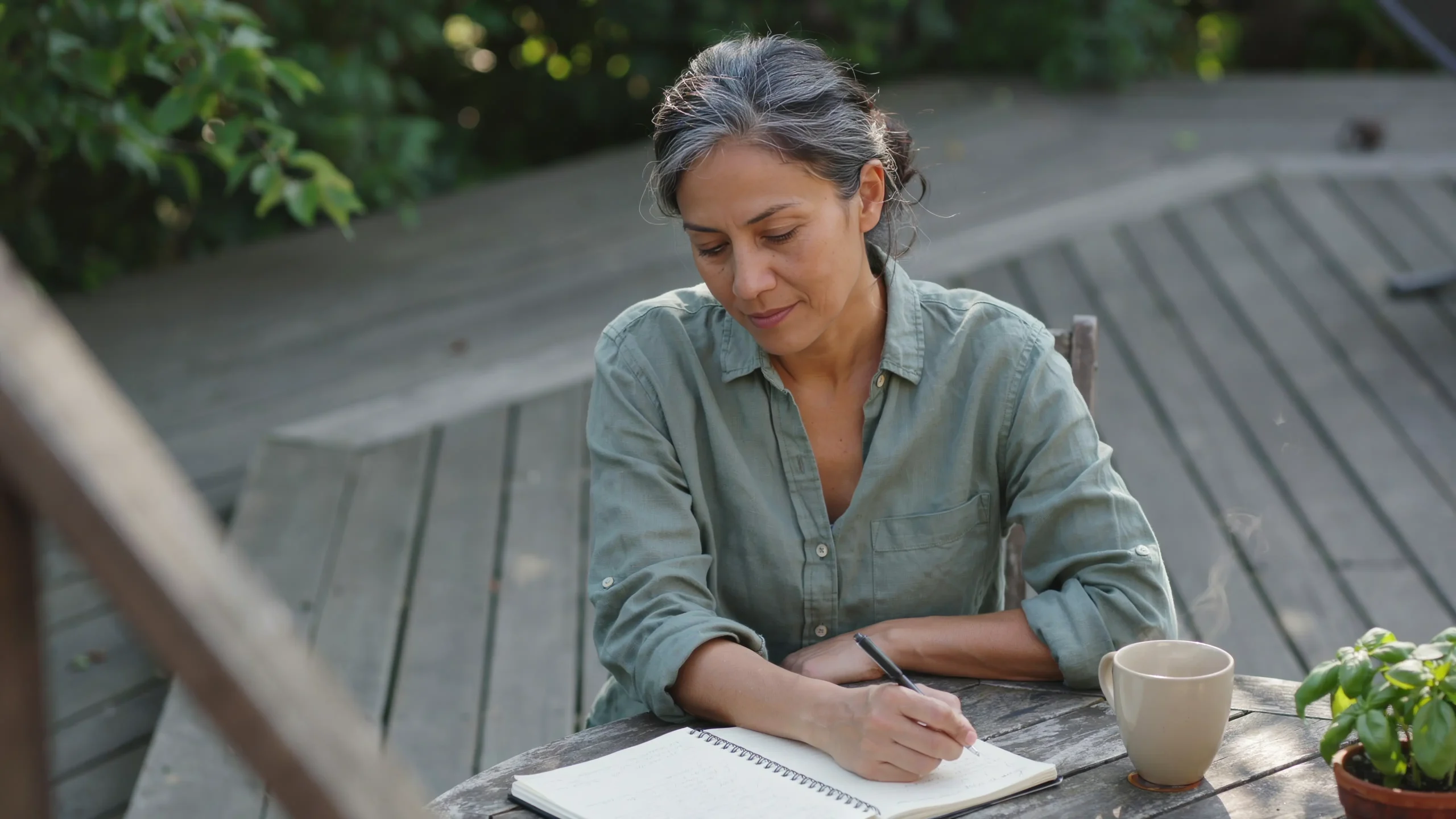 A Latina woman in her mid-50s sits at an outdoor table with an open notebook and coffee, looking thoughtfully at her writing on a quiet summer morning — the quiet work of building what's next without a blueprint.