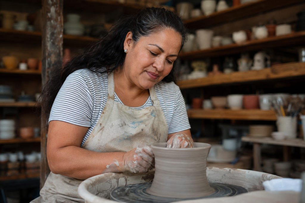 A woman shapes wet clay at a pottery wheel in a community studio, trying something new in the exploring stage of reinvention.